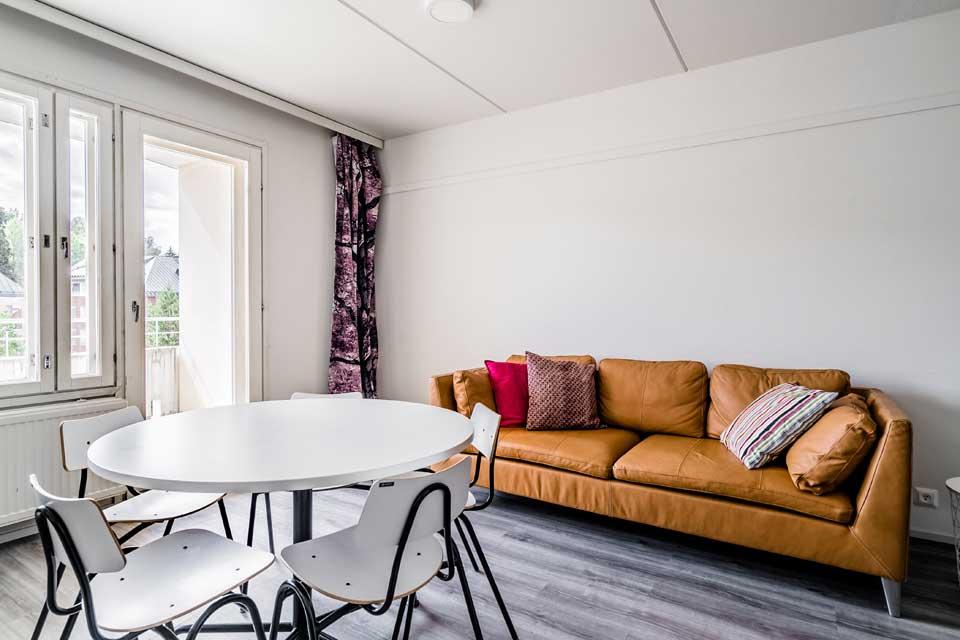 White dining table with four chairs in front of the picture. Behind the table, against the wall is an brown leather couch with colorful throw pillows.  Glass door to the balcony next to the couch. Room has white walls and grey wood-like floor.